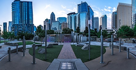 Panoramic view of a modern urban park surrounded by city skyscrapers at dusk. The park, with its diverse greenery, stone paths, and metallic sculptures, offers a serene escape. The tall buildings in the background reflect the evening light. Nearby, new apartment barn kits blend seamlessly into the picturesque scene.