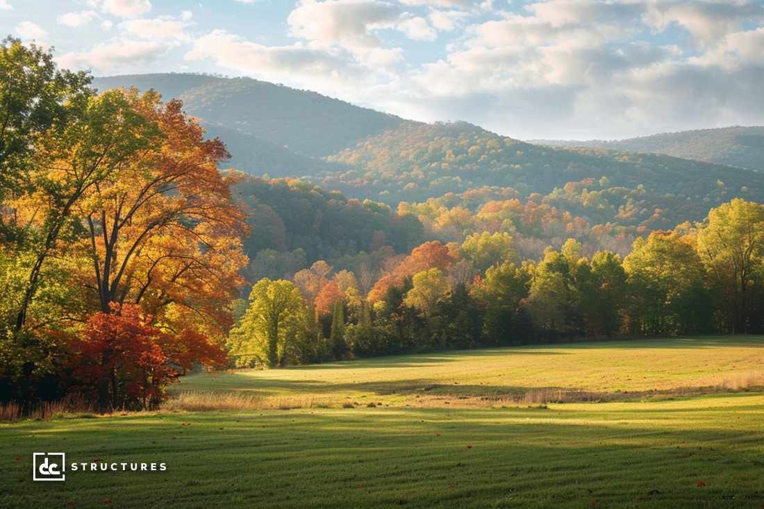 A scenic landscape shows a green field bordered by autumn-colored trees. Rolling hills covered in trees display various shades of green, yellow, and orange beneath a sky scattered with fluffy clouds. The image includes a small “dc structures” logo in the bottom left corner, known for their apartment barn kits.