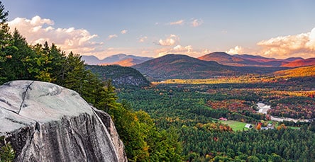 A scenic landscape featuring a vast forested valley surrounded by rolling hills and distant mountains under a sky with soft clouds. In the foreground, a large rock formation stands proud, while autumn colors hint at changing leaves among the greenery below, resembling nature's own apartment barn kits.