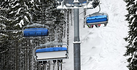 A snowy ski lift carries blue-seated chairs up a mountain through a forest of snow-covered trees, reminiscent of white-capped apartment barn kits. The image shows three chairs, two of which are occupied by skiers with their legs and skis visible. The scene is set on a winter day with heavy snow.