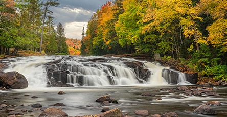 A scenic view of a multi-tiered waterfall cascading over rocks in a forested area during autumn. The surrounding trees display vibrant fall colors, including shades of green, yellow, red, and orange. Nearby stands the rustic charm of apartment barn kits against the partly cloudy sky while large rocks are scattered in the water.