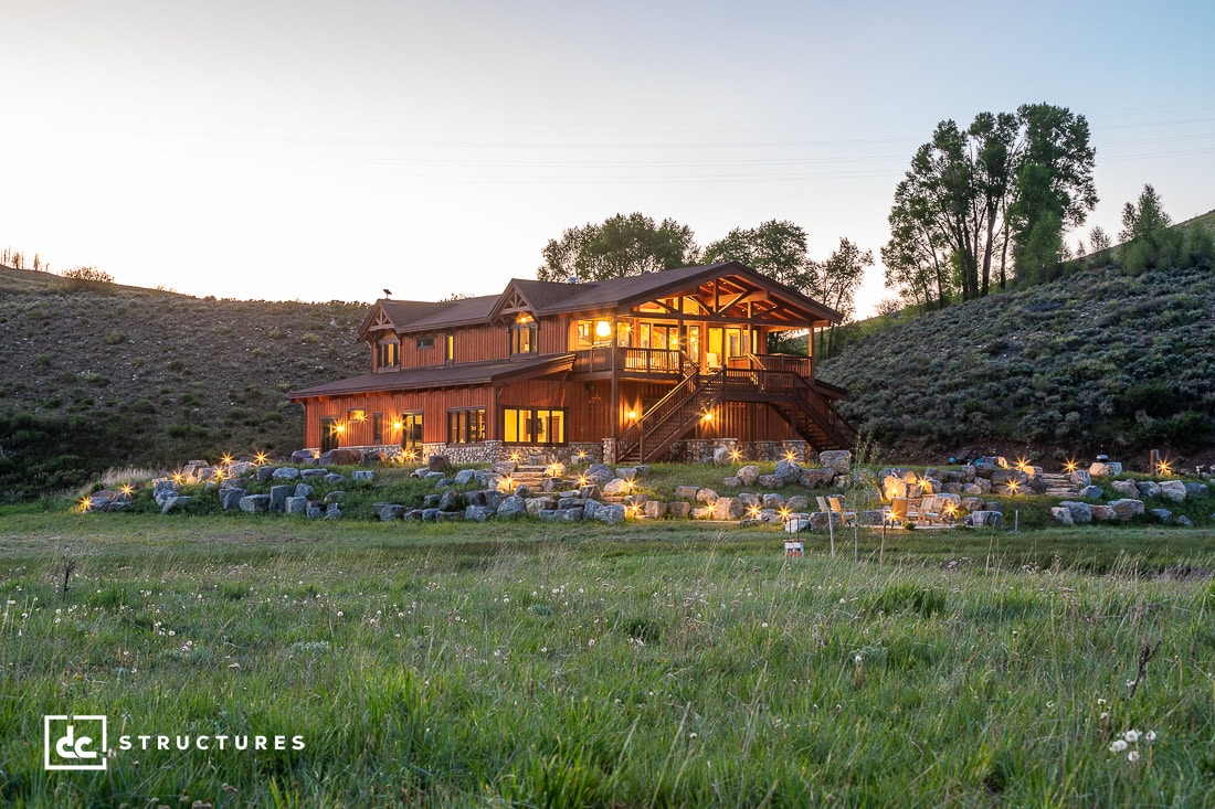 A large, rustic wooden house with multiple peaked roofs is illuminated warmly at dusk. The house sits on a gently sloping hill surrounded by grass and scattered rocks. A logo in the bottom left corner reads "dc structures.