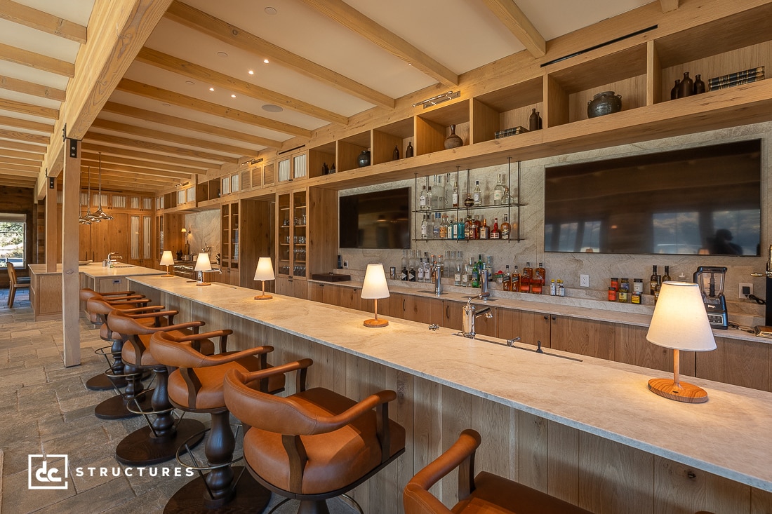 A wide-angle view of a stylish bar interior with a long marble countertop lined with wooden bar stools. The bar is well-lit with modern pendant lights, featuring multiple liquor bottles, glassware, and two large flat-screen TVs mounted on the wooden shelves.