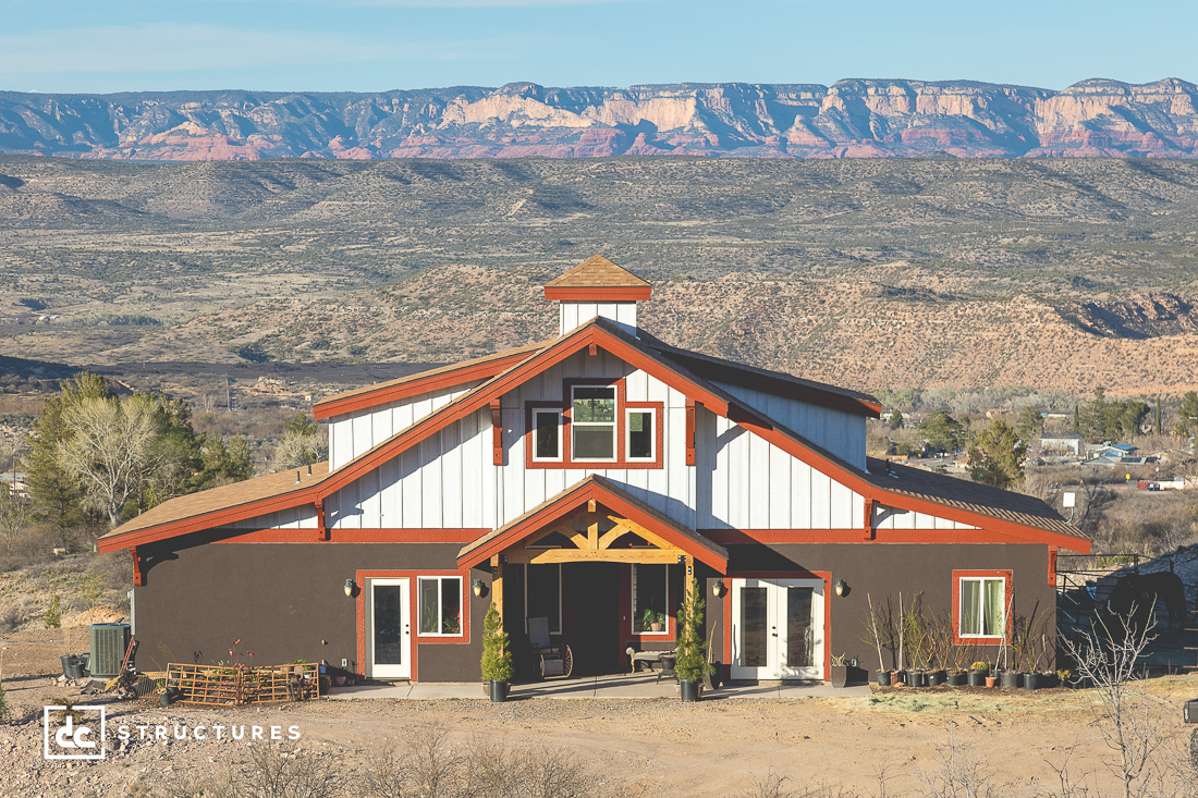 A large barn-style house with white and brown exterior stands in a rural area with mountains in the background. The house features a central peaked roof, multiple windows, and a welcoming front porch. The landscape includes dry terrain and sparse vegetation.