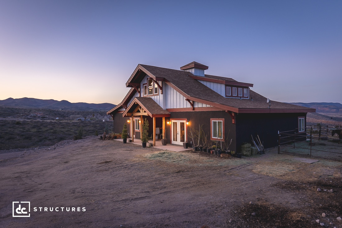 A rustic house with large wooden beams and dark exterior set against a sunset backdrop. The house features a gabled roof, dormer windows, and a small porch. The surrounding landscape consists of rolling hills and open land. The logo "dc Structures" is visible in the corner.