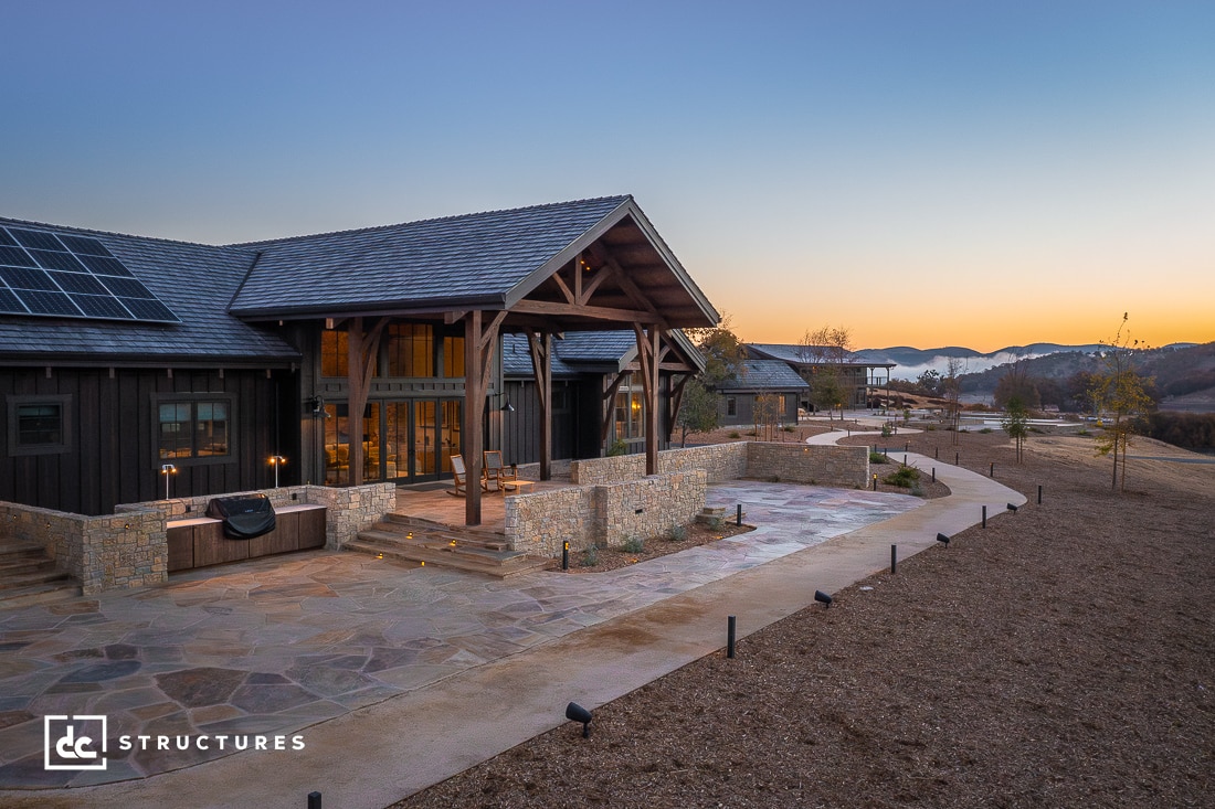 An elegant, modern barn-style home at sunset, surrounded by scenic landscapes. The building features large windows, stone accents, and wooden beams. Solar panels are visible on the roof, and a spacious patio with pathways extends into the yard.