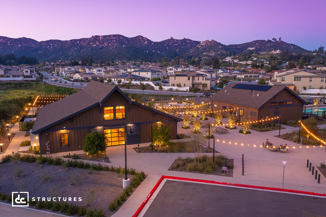 An aerial view of a residential area at sunset, featuring two barn-style buildings surrounded by string lights and pathways. The scene is set against a backdrop of hilly terrain and scattered houses, with the landscape bathed in a soft, violet twilight.