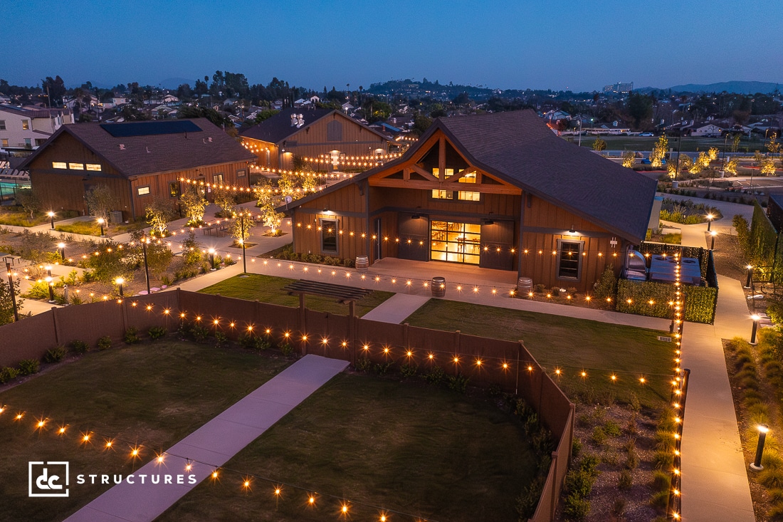 Aerial view of a modern barn-style building lit up with string lights at dusk. The building, with large windows and wooden beams, is surrounded by landscaped paths and greenery. The scene is serene with a soft, warm glow over the entire area. Logos "dc Structures".