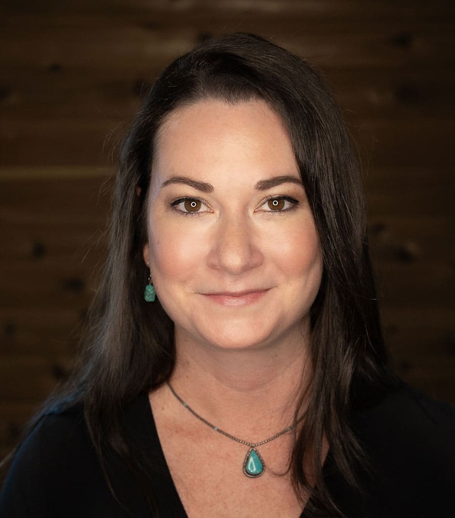 Portrait of a woman with long, dark hair and light skin, wearing a black top. She has a necklace with a teardrop-shaped turquoise pendant and matching earrings. She smiles softly and the background is a blurred, wooden texture.