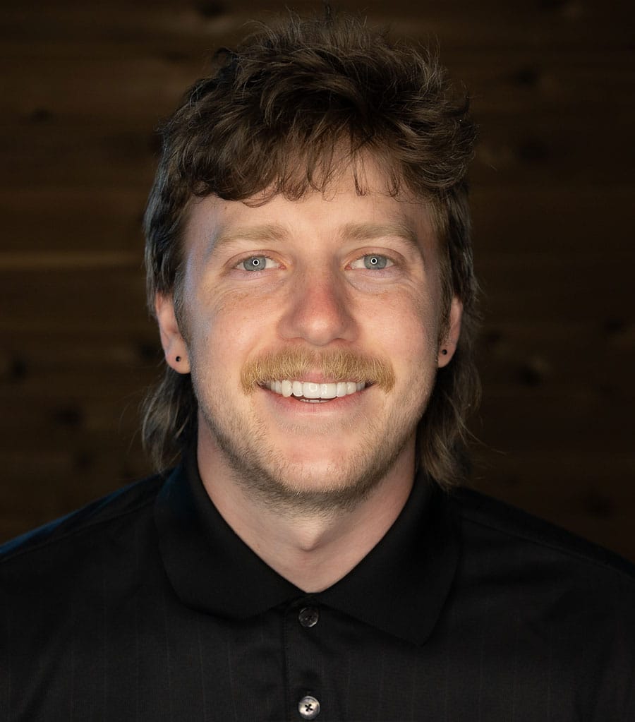 A man with light brown hair and a mustache is smiling at the camera. He is wearing a black collared shirt, and the background is made of dark wooden panels.