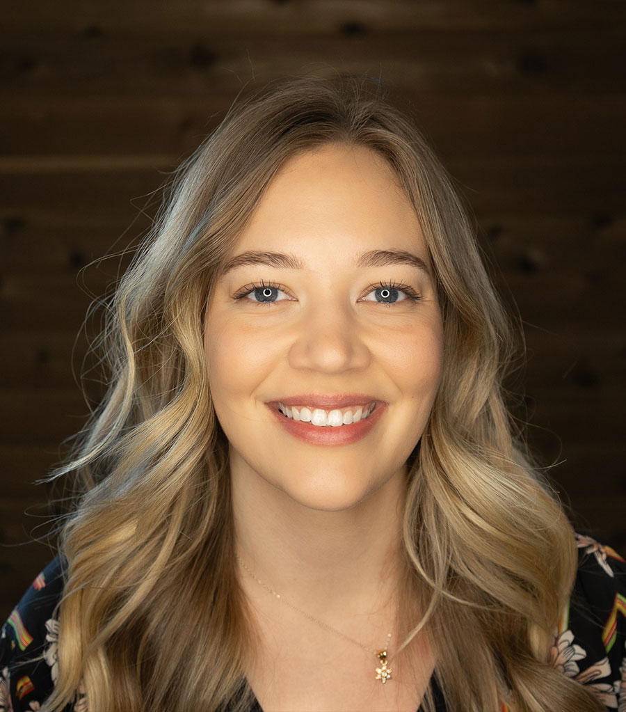 A young woman with long, wavy blonde hair and blue eyes smiles at the camera. She is wearing a dark top with a colorful pattern and a delicate necklace with a small pendant. The background is a dark wooden wall.