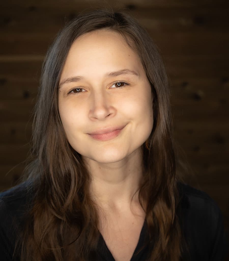 A person with long, brown hair and a slight smile looks directly at the camera. They are wearing a dark-colored shirt, and the background is softly illuminated with a wooden texture.