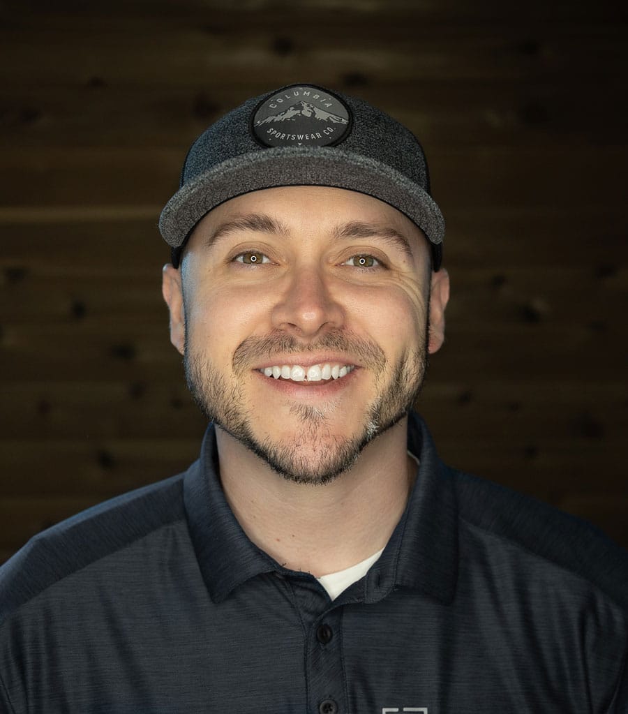 A man with a friendly smile is wearing a dark baseball cap with a logo and a dark collared shirt, standing in front of a wooden background.