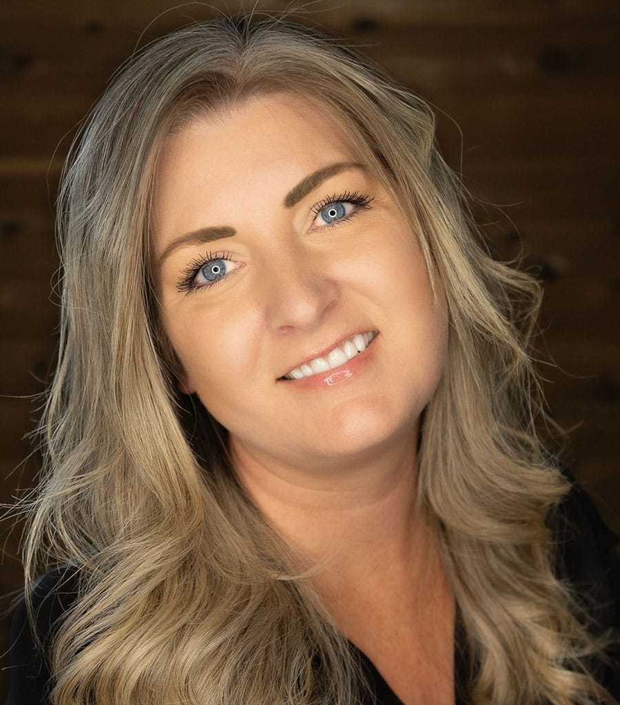 A woman with long, wavy blonde hair smiles at the camera. She has blue eyes and is wearing natural makeup. The background is blurry and dark, which makes her face stand out clearly.