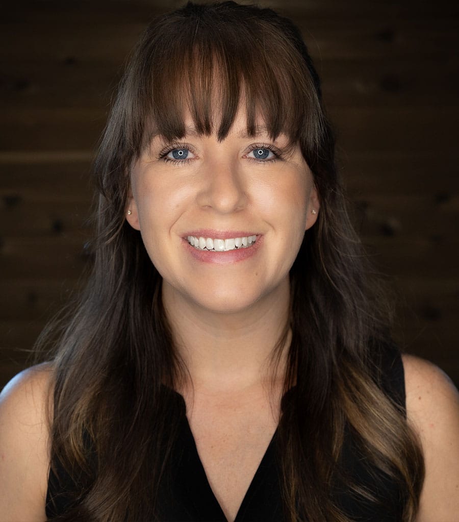 A woman with long brown hair and bangs is smiling brightly at the camera. She is wearing a sleeveless black top and has blue eyes. The background is dark wood, giving a warm and rustic feel to the image.