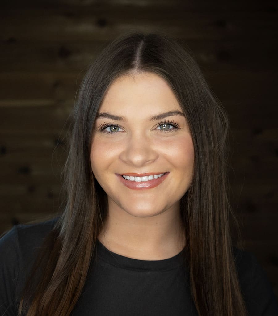 A woman with long, straight brown hair and blue eyes is smiling at the camera. She is wearing a black top. The background is a dark wooden wall.