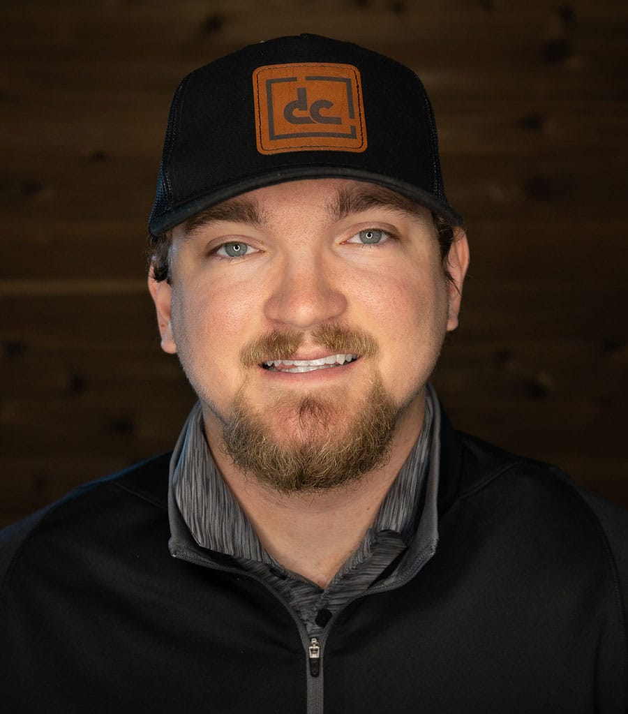 A man with a beard and mustache is smiling at the camera. He is wearing a black baseball cap with a brown rectangular logo, a black jacket, and a gray shirt. The background is a wooden wall.