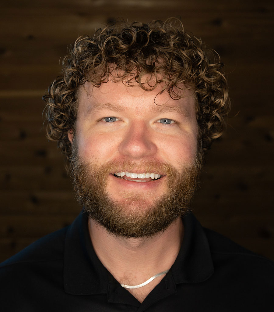 A person with curly hair, a beard, and a mustache smiles at the camera. They are wearing a black shirt, and the background is a dark, blurred surface with a wood-like texture.