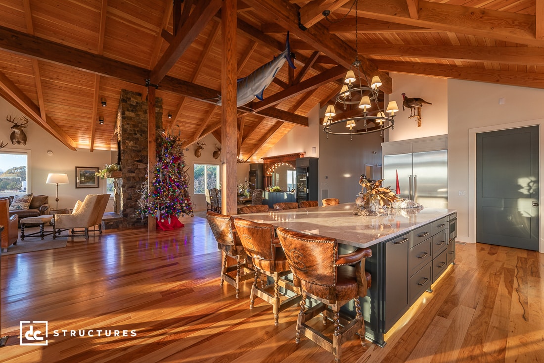 A spacious living area with a high wooden ceiling, featuring a large kitchen island with leather chairs. A decorated Christmas tree stands near a stone fireplace, and deer heads are mounted on the walls. Sunlight streams through the windows, illuminating the space.