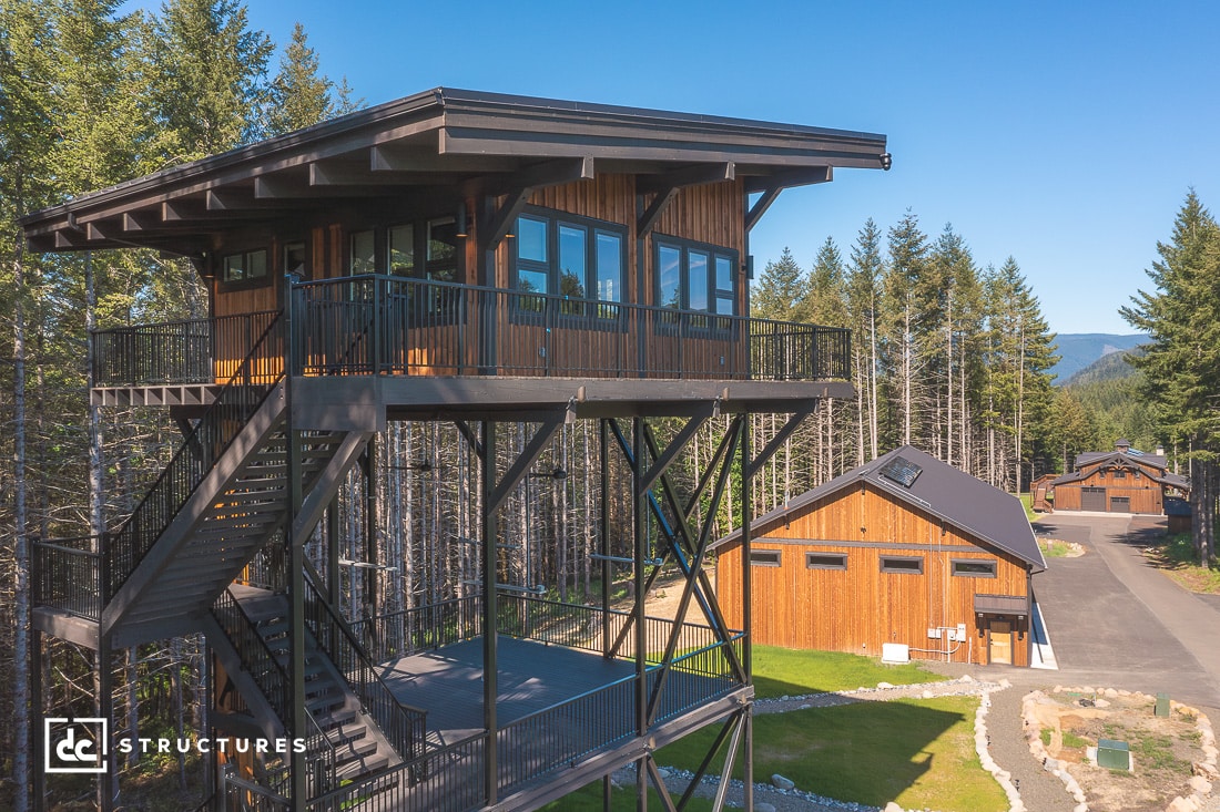 A modern treehouse-style structure elevated on metal supports with an outdoor staircase. It overlooks a wooden building in a wooded area, surrounded by tall trees and mountains in the background.