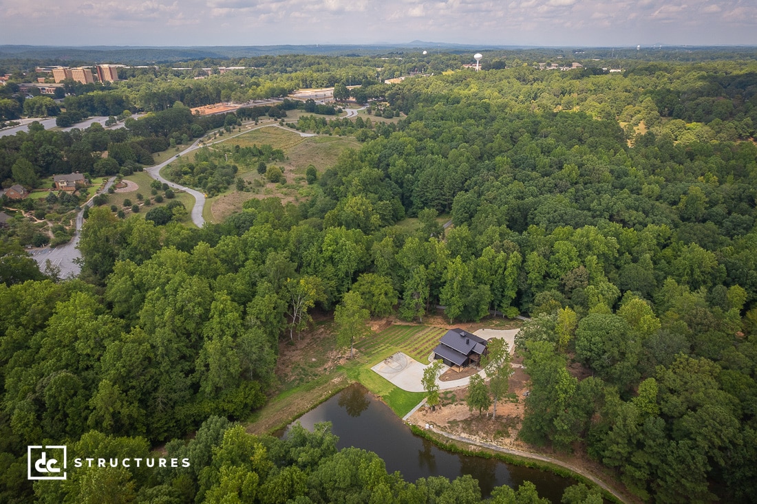 Aerial view of a wooded area with a small pond and a modern wooden house. Roads and scattered buildings are visible in the distance. The landscape is lush with greenery under a partly cloudy sky.