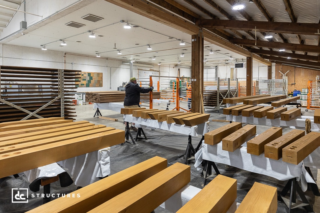 A person works in a spacious industrial workshop with stacked wooden beams, reminiscent of apartment barn kits. The room features overhead lighting and exposed beams, with various tools and materials neatly organized around the space.