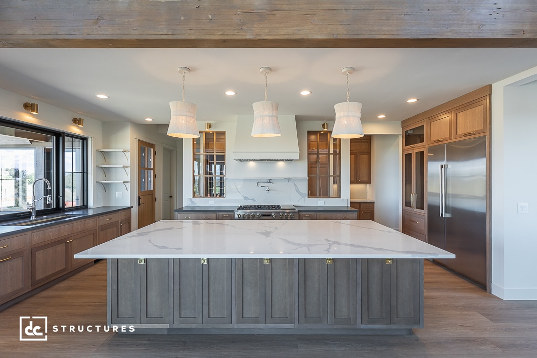 Modern kitchen with a large marble-topped island, featuring wooden cabinetry and stainless steel appliances typical of stylish barndominium kits. Three pendant lights illuminate the space, which includes a double-door refrigerator and gas stove. Natural light pours in through windows on the left.