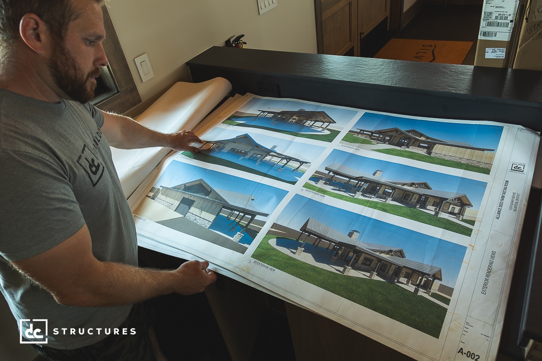 A man examines architectural blueprints showcasing different views of an apartment building design. The office, with its wooden interior, adds warmth as the plans for barn kits are spread out on a dark table. A logo says 