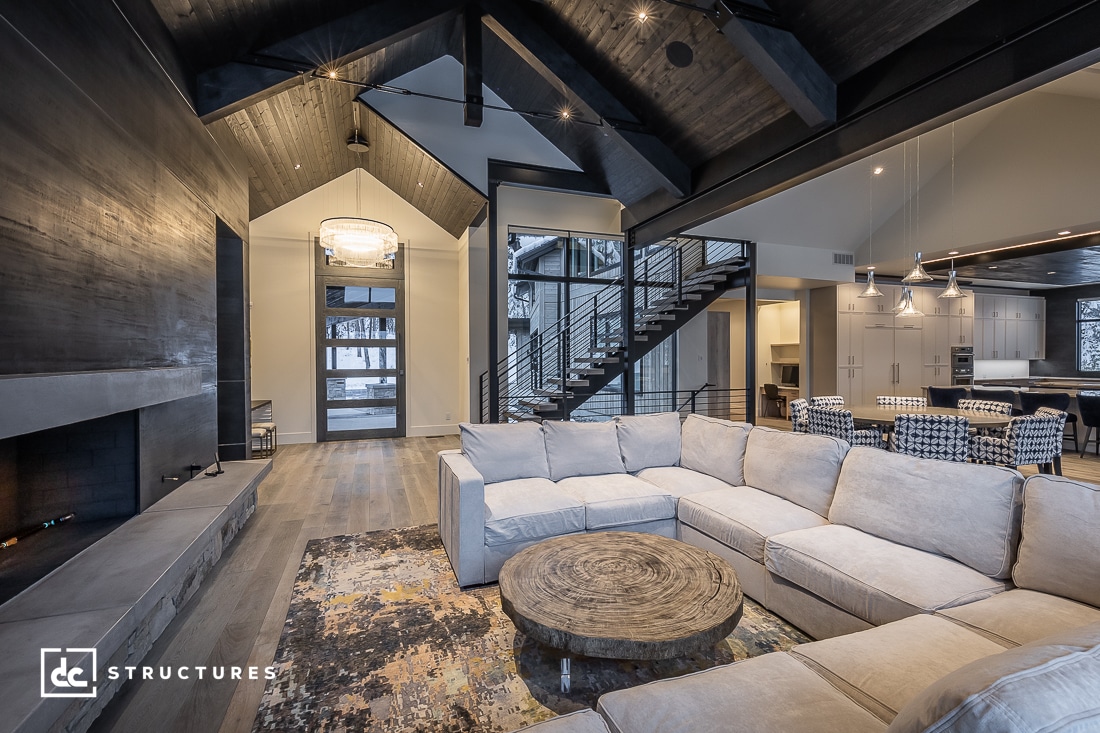 Spacious modern living room featuring elements from modern home kits, with a large beige sectional sofa, rustic round coffee table, high vaulted dark wood ceiling, and an open kitchen area with hanging pendant lights. Stairs lead to an upper level, and a large window offers natural light.