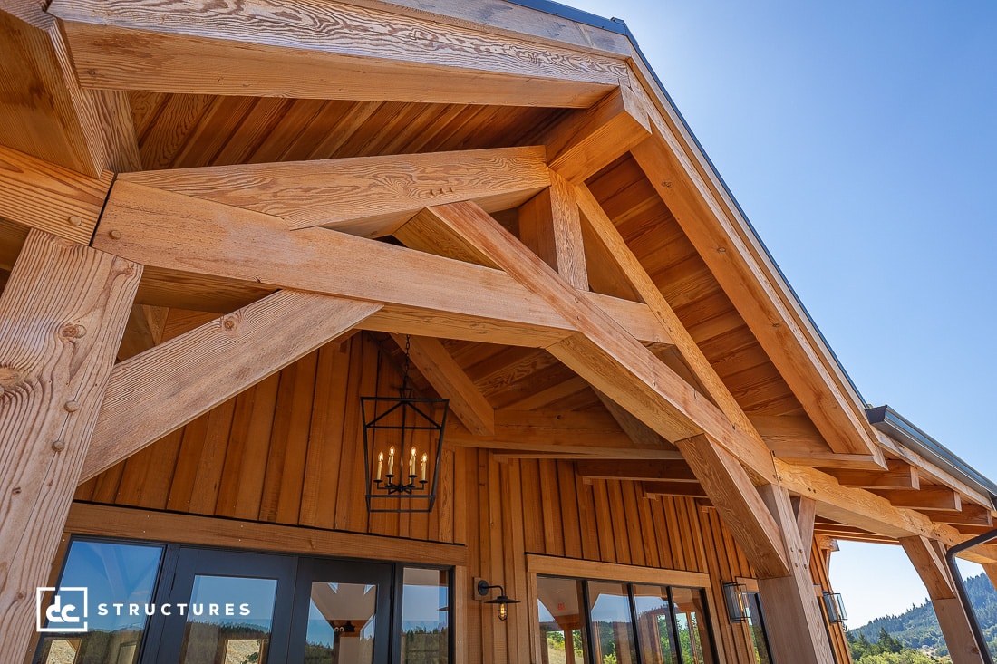 Close-up of a rustic timber frame home kit, the wooden building exterior features exposed beams and a triangular roof. A black metal lantern with candles hangs near the entrance, enhancing its charm. The sky is clear and blue in the background.