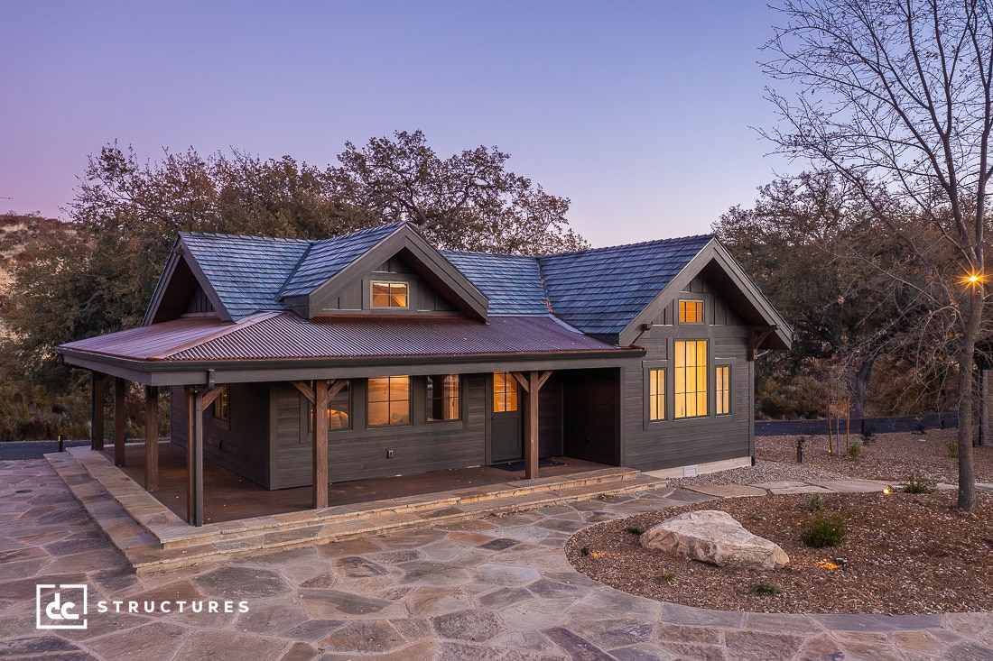 A cozy, dark wood cabin kit with a wraparound porch stands amidst a natural setting at dusk. Warm light glows from the windows, and a stone pathway leads to the entrance. Bare trees and a hilly backdrop complete this tranquil, ready-to-assemble scene.