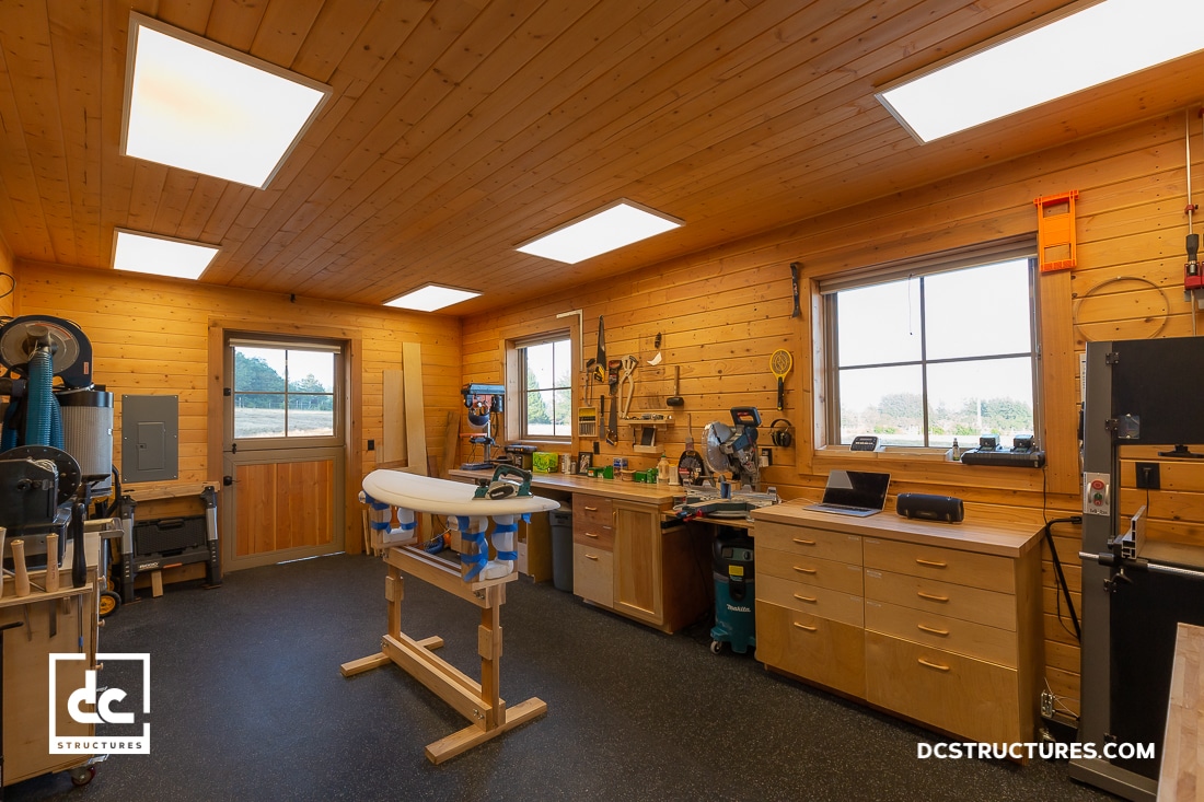 A well-organized woodworking workshop featuring wooden walls and ceiling, bright overhead lighting, and large windows. The room is equipped with a central workbench, various tools, garage kits for assembly projects, and ample storage cabinets.