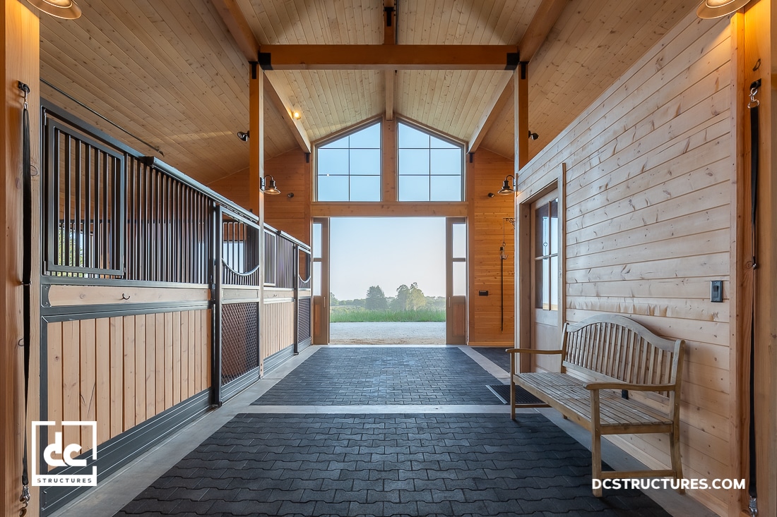 A spacious, wooden horse stable interior with high ceilings and large windows letting in natural light, reminiscent of our garage kits. The stable features a bench on the right side and a view of greenery through the open doors. The DC Structures logo is visible at the bottom.