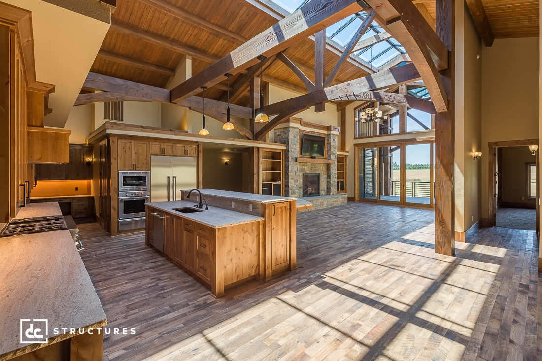 Spacious rustic kitchen with wooden beams and high ceilings in a barn home kit style. Large windows illuminate the central island, equipped with a sink, oven, and wooden cabinets. Sunlight streams through skylights, enhancing the wood-paneled floors and walls.