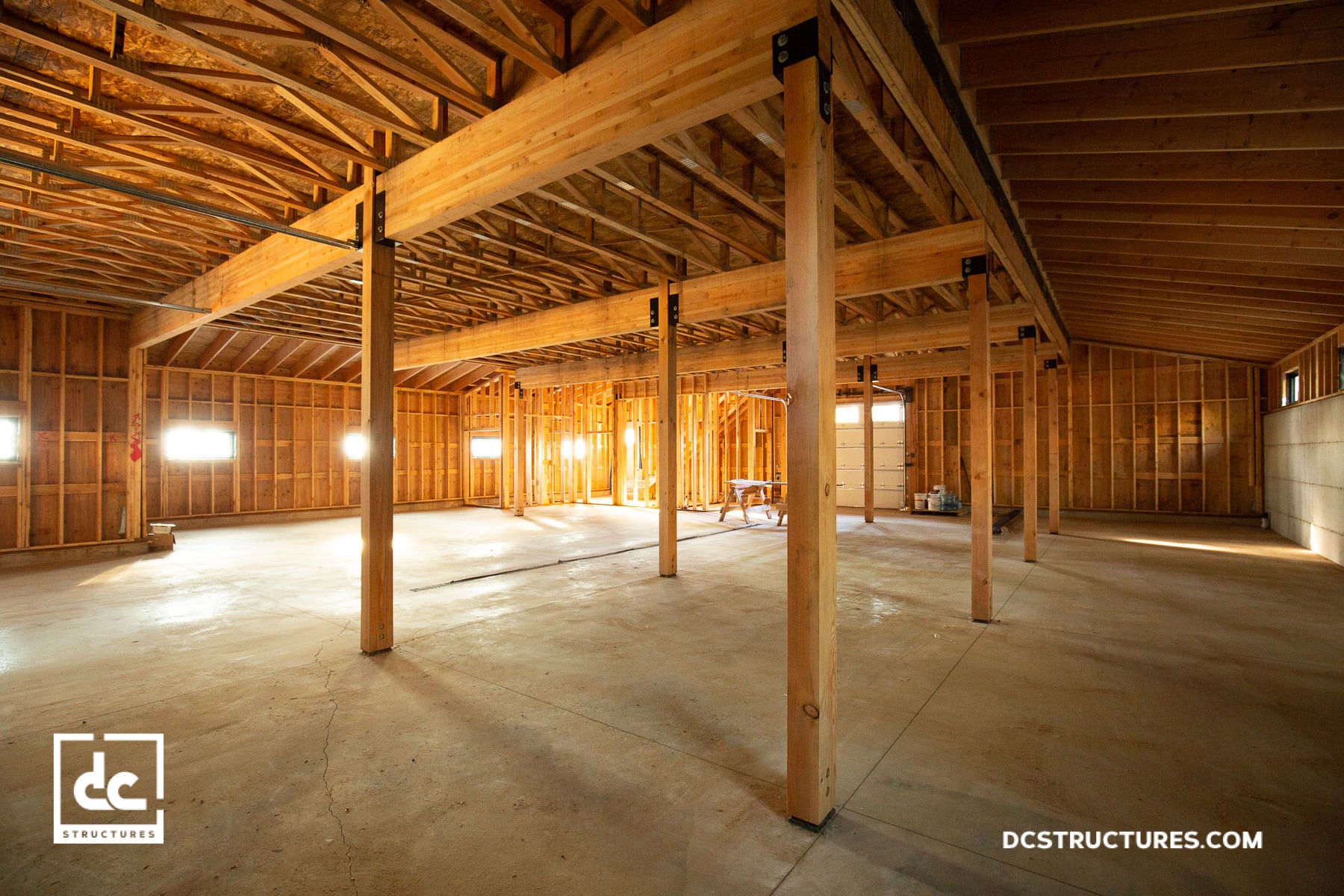 Interior of a spacious, unfinished wooden structure with exposed beams and a concrete floor, akin to barn home kits. Sunlight streams in through windows, illuminating the open space with warmth. In the background, construction materials hint at future possibilities.