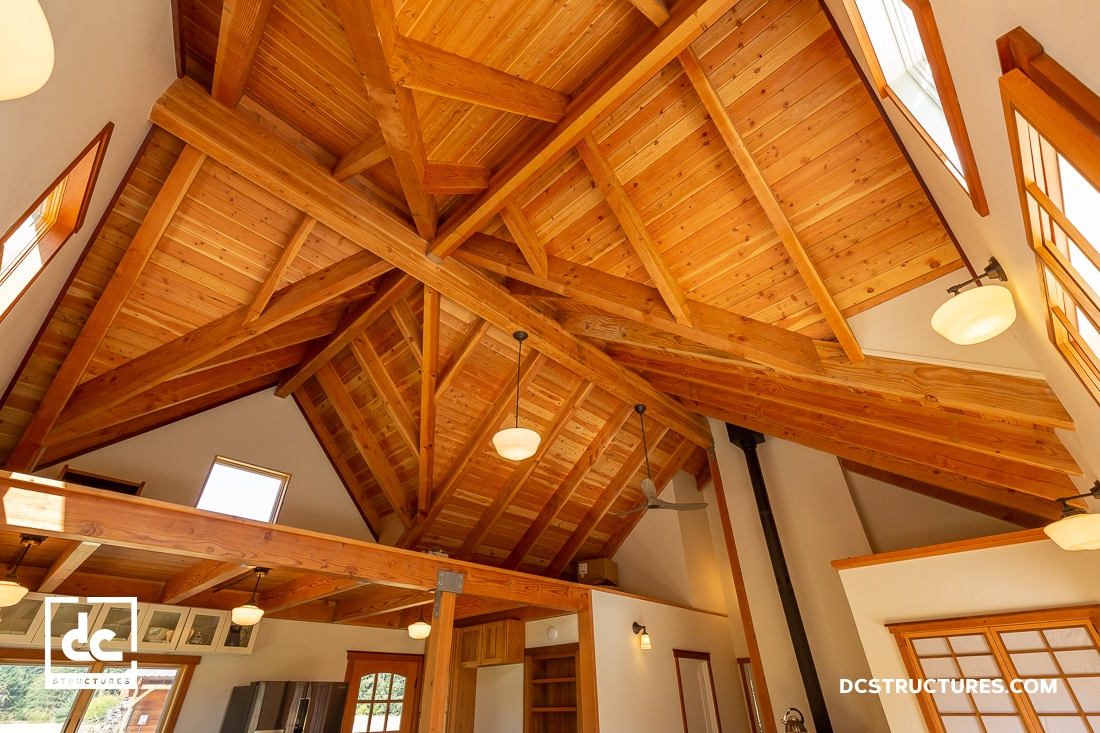 A spacious interior with a high wooden ceiling featuring exposed beams and warm lighting. Inspired by cabin kits, the loft area is visible, and large windows allow natural light to illuminate the space. The design emphasizes natural wood and open architecture.