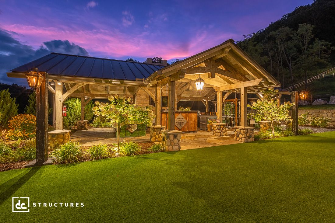 A beautifully lit outdoor pavilion at dusk, crafted from premium pavilion kits, features a wooden frame, metal roof, and stone accents. The manicured lawn and trees add to the serene ambiance as the sky transitions from pink to deep purple tones in the background.