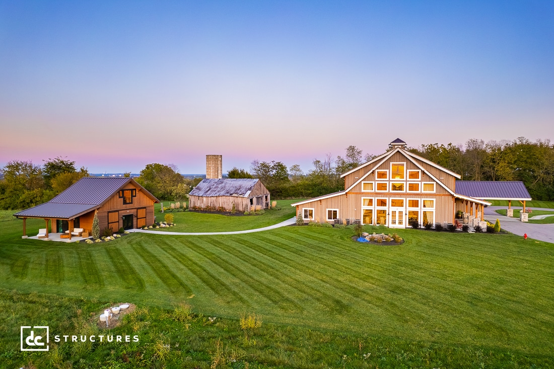 A picturesque rural scene at sunset showcases a large barn-style building, reminiscent of popular wedding barn kits, with numerous windows, a silo, and a charming wooden structure. The well-manicured lawn stretches out front as trees gently sway in the background.