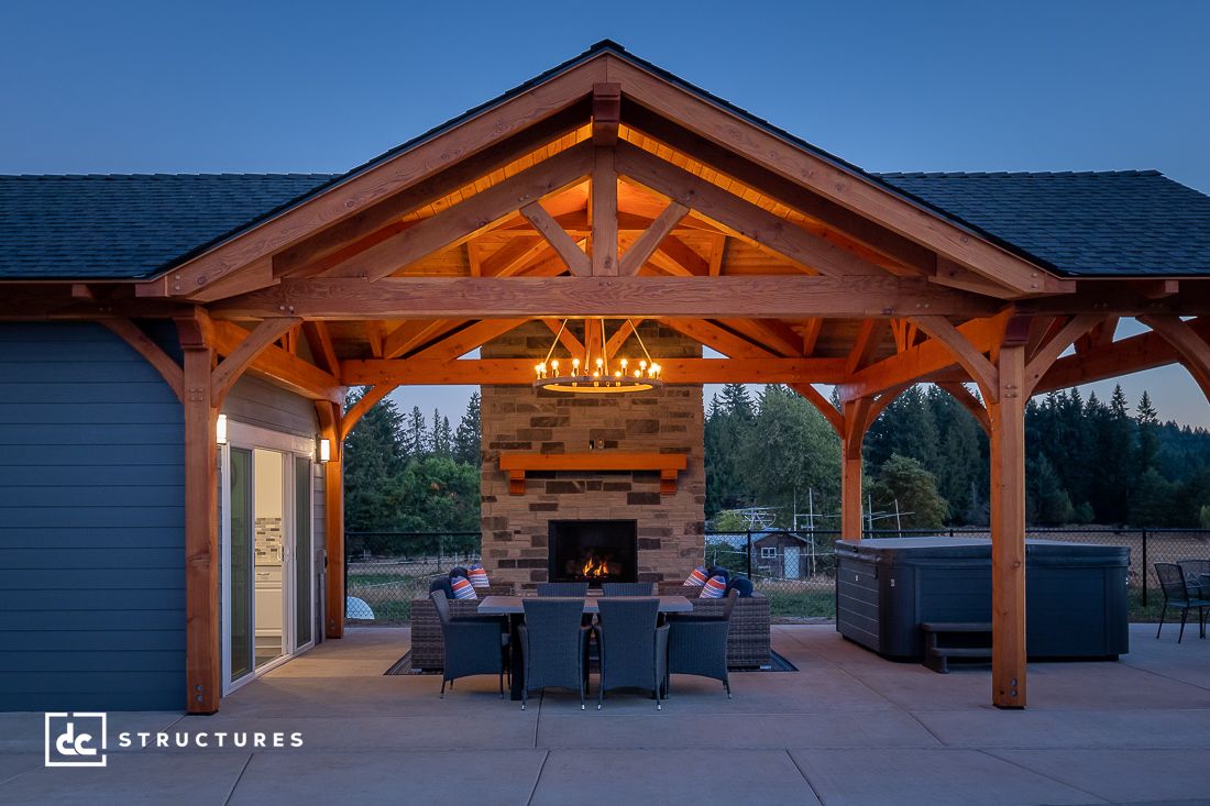 A wooden pavilion kit with a gabled roof features a stone fireplace and a chandelier above the dining table set. It opens to a well-lit outdoor space with greenery in the background, under a tranquil dusk sky.