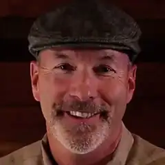 A smiling man with a goatee and mustache wears a flat cap and a beige shirt. The background is dark brown, suggesting a cozy indoor setting.