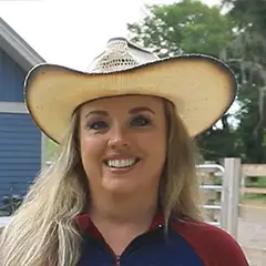 A smiling woman wearing a large cowboy hat stands outside. She has long blonde hair and is dressed in a red and blue top. In the background, there are trees and a blue building.