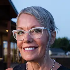 A smiling woman with short gray hair and white-framed glasses is wearing a necklace and a black top. She is standing outside with a blurred outdoor structure and sky in the background.