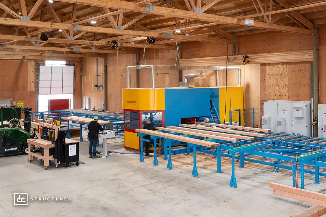 The interior of a spacious woodworking shop features a blue and yellow automated cutting machine, ideal for crafting apartment barn kits. Workers skillfully operate the equipment as wooden beams glide on conveyor belts. The shop boasts wooden walls, high ceilings, and abundant natural light from large windows and doors.