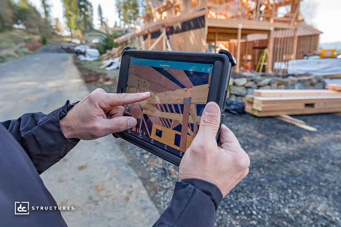 A person holds a tablet displaying a digital model of building beams while pointing at it. In the background, a partially constructed barn home kit is visible, featuring wooden beams and some construction materials scattered on the ground.