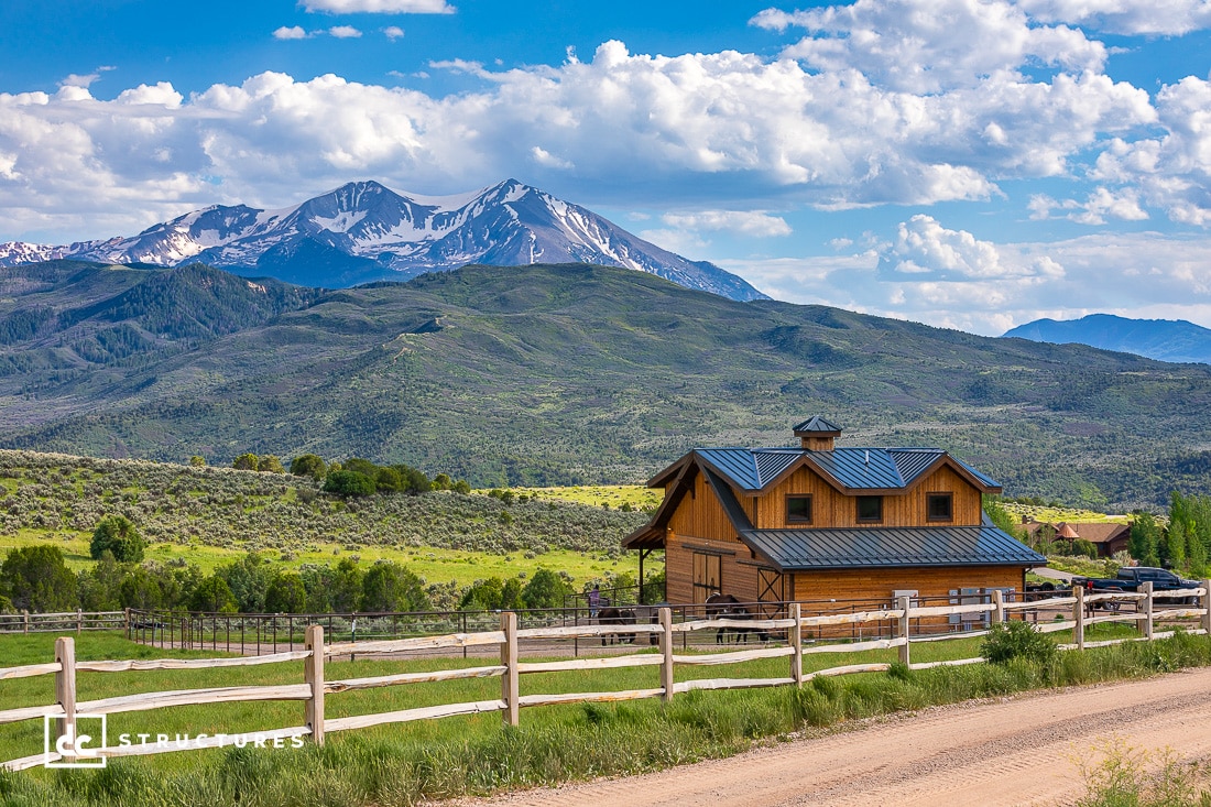 A rustic wooden barn with a blue roof sits on a rural dirt road, surrounded by lush green hills. In the background, snow-capped mountains rise under a partly cloudy sky. A wooden fence lines the foreground.