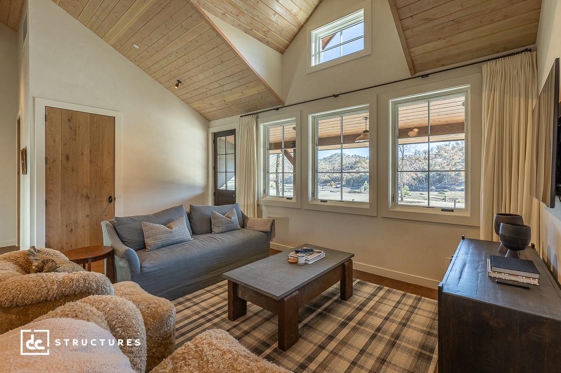 A cozy living room in an apartment barn with a wooden ceiling and large windows. It features a gray sofa, two plush armchairs, a wooden coffee table, and a dark wooden TV stand. A plaid rug covers the floor as sunlight streams through the windows.