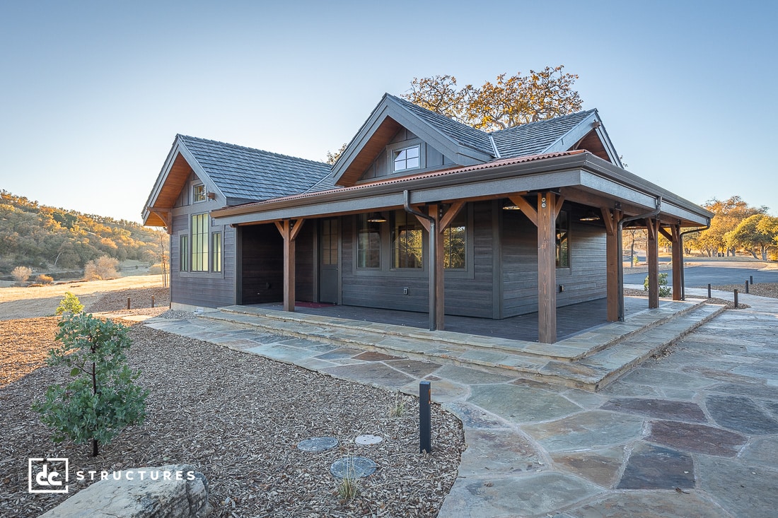 A modern cabin with a wooden exterior, reminiscent of apartment barn kits, is surrounded by a rustic stone patio. The house features a sloped roof and large windows. The dry landscape has scattered trees in the background beneath a clear sky.