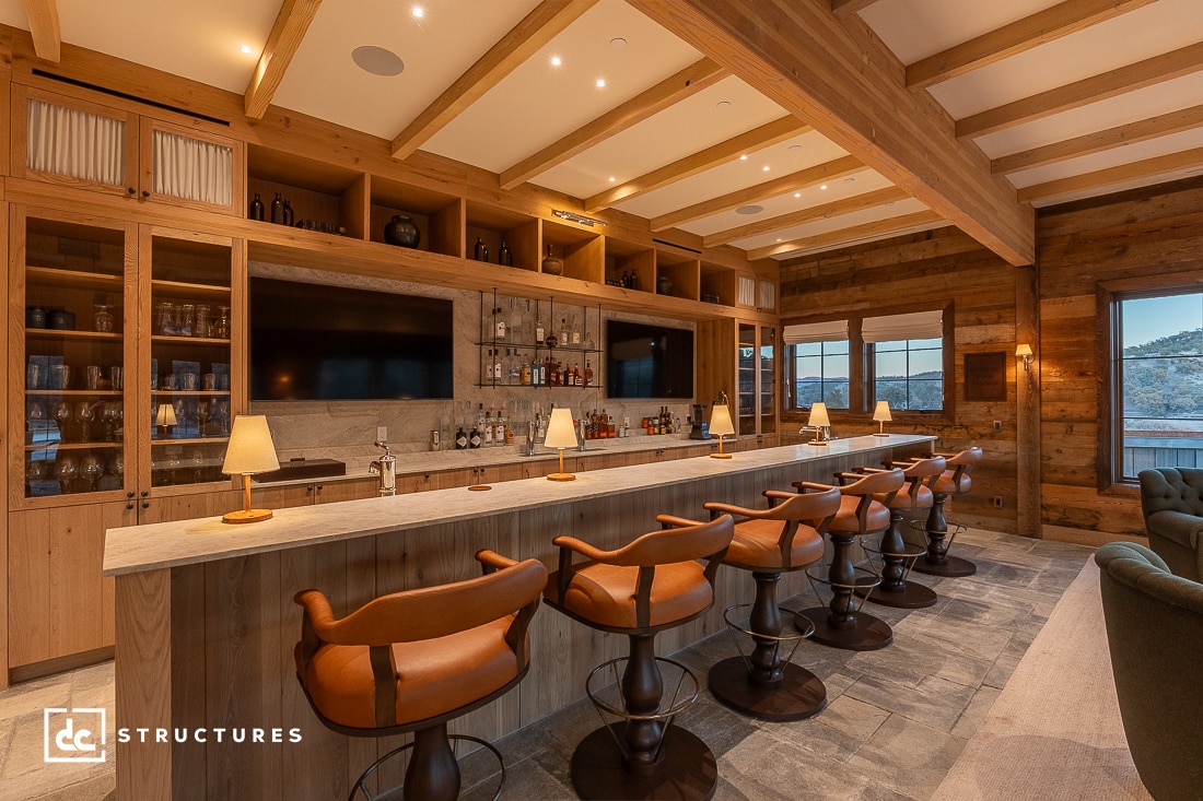 A cozy bar interior with wooden beams and shelves. The bar counter is lined with six brown leather stools, and dim lighting creates a warm ambiance. Shelves behind the counter display bottles and decor. Large windows provide an outside view.