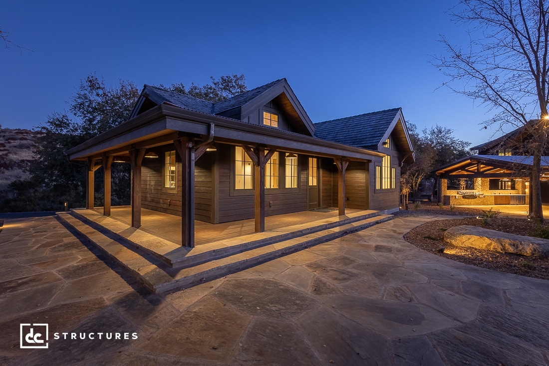 A cozy, modern cabin crafted from apartment barn kits features a wraparound porch illuminated at dusk. The structure, made of dark wood, is surrounded by a stone patio and bare trees under a clear evening sky.