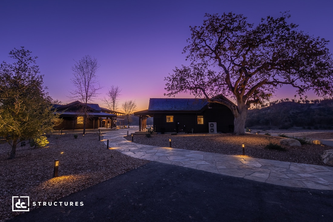 A scenic dusk view of modern cabins with a stone path, surrounded by trees. The sky displays a vibrant purple and orange gradient. Ground lights illuminate the path, creating a serene and inviting atmosphere. A large tree stands prominently to the right, reminiscent of rustic apartment barn kits in design.